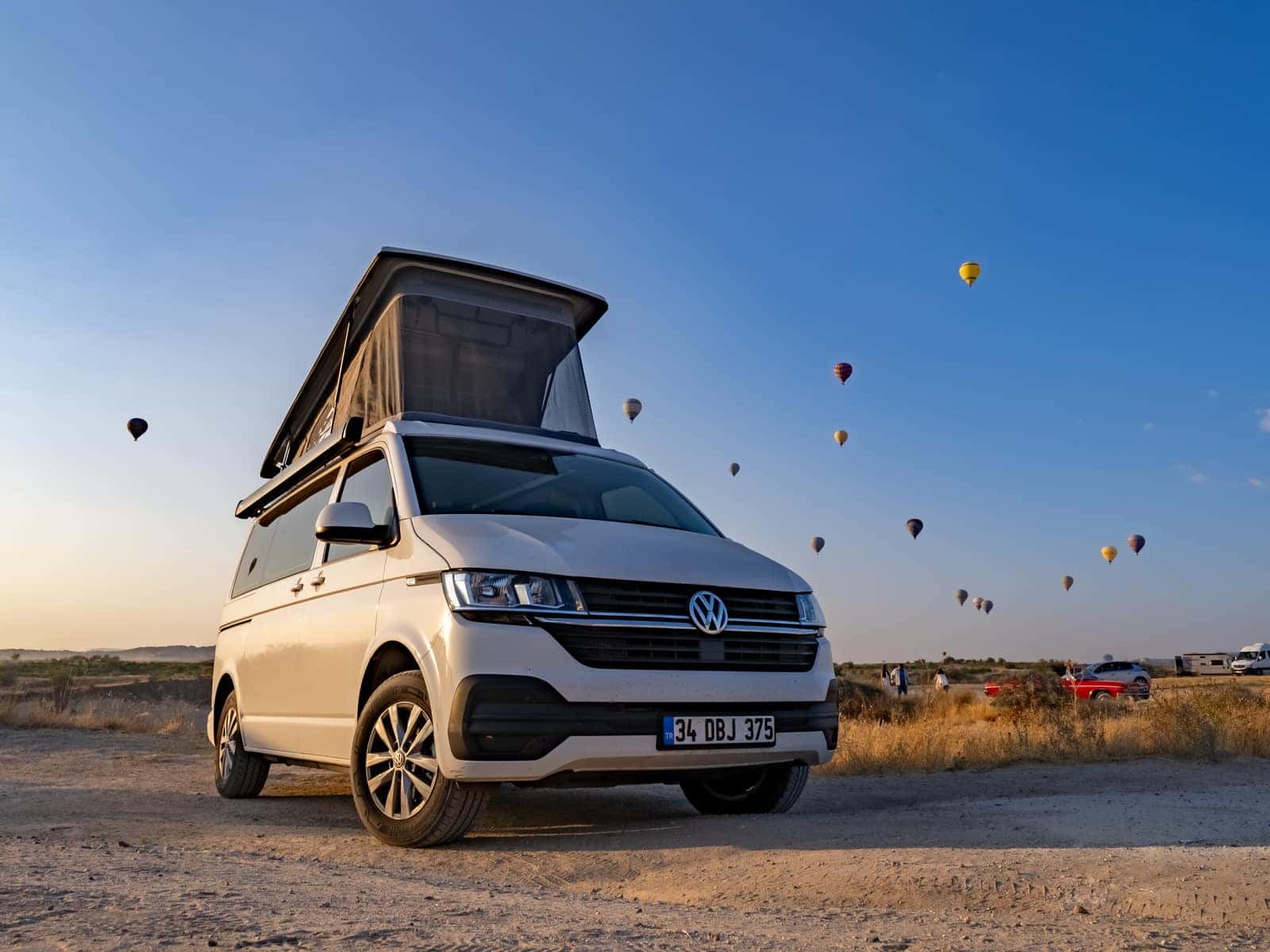 Beyaz VW T6.1 tabanlı Cappadocia, gün doğumunda Kapadokya ovasında; pop-up tavanı açık, gökyüzünde sıcak hava balonları