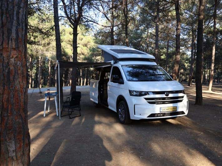 Beyaz yeni nesil VW Transporter uzun şasi üzerine kurulu Ephesus, çam ormanında; pop-up tavanı ve tentesi açık