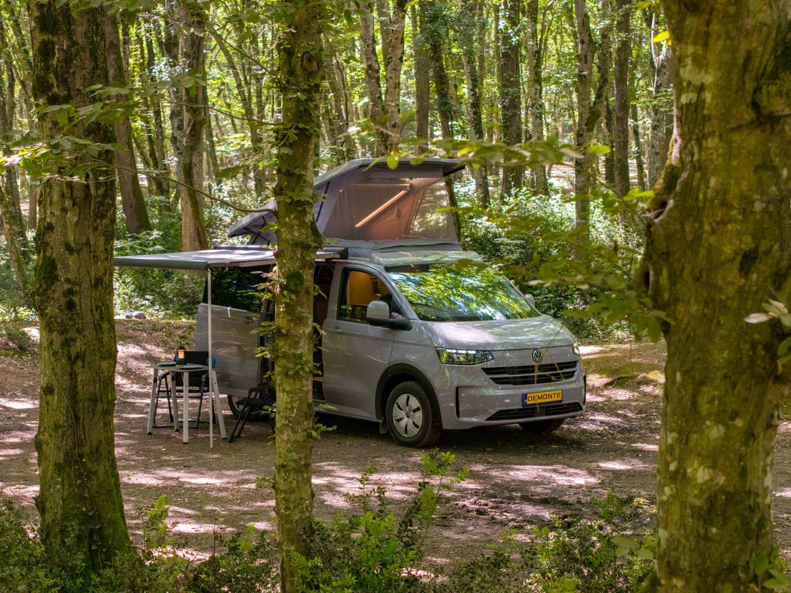 Yeni nesil VW Transporter tabanlı New Cappadocia ormanda; pop-up tavanı ve tentesi açık, altında kamp masası